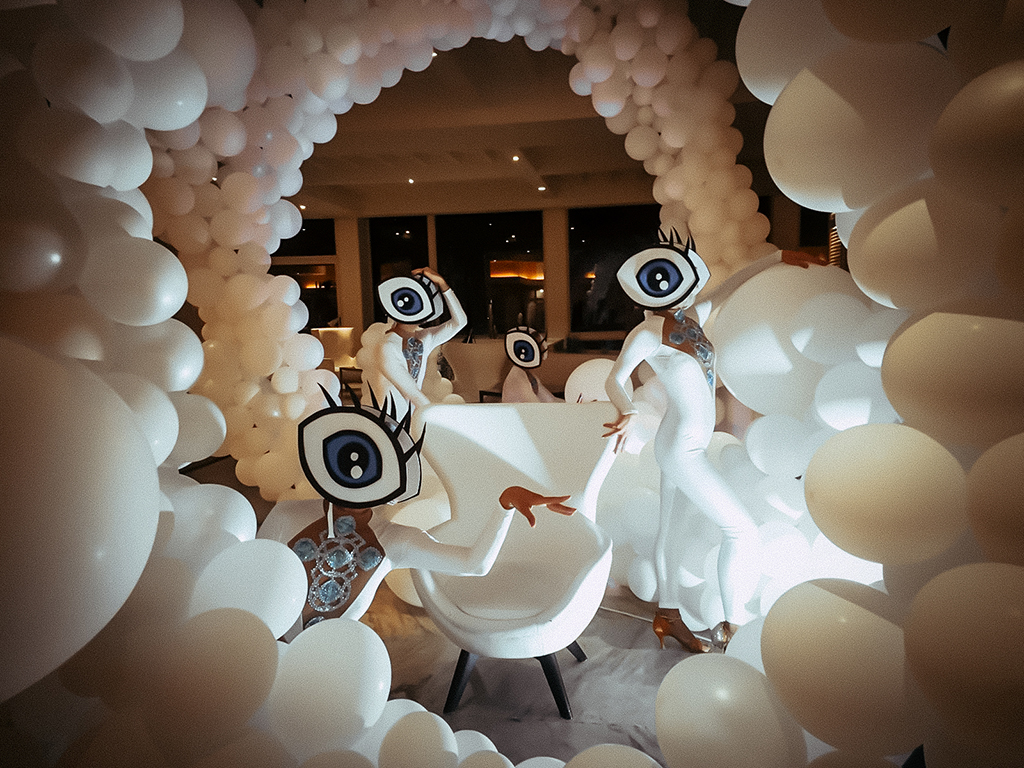 Mensen in gigantische oogkostuums omringd door witte ballonnen in een kamer.
