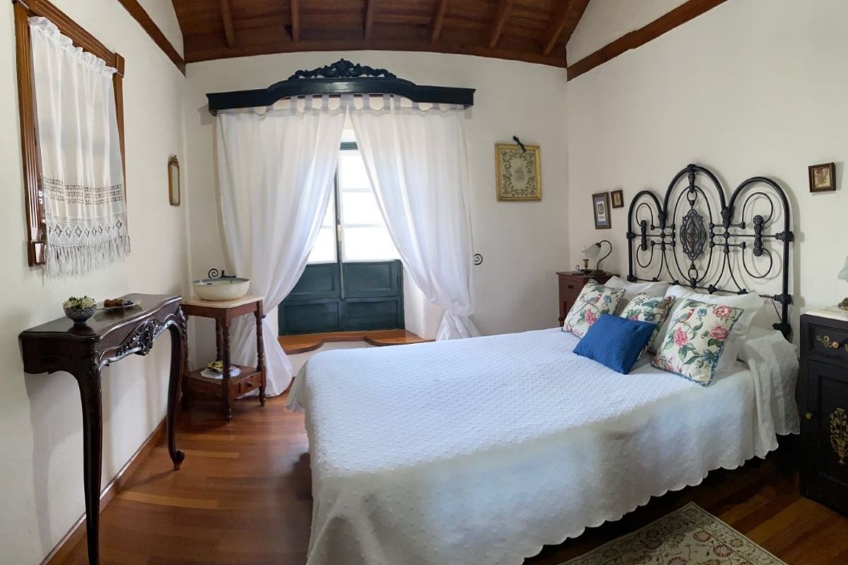 Kamer met smeedijzeren bed, bloemkussens en raam met witte gordijnen in Tenerife.