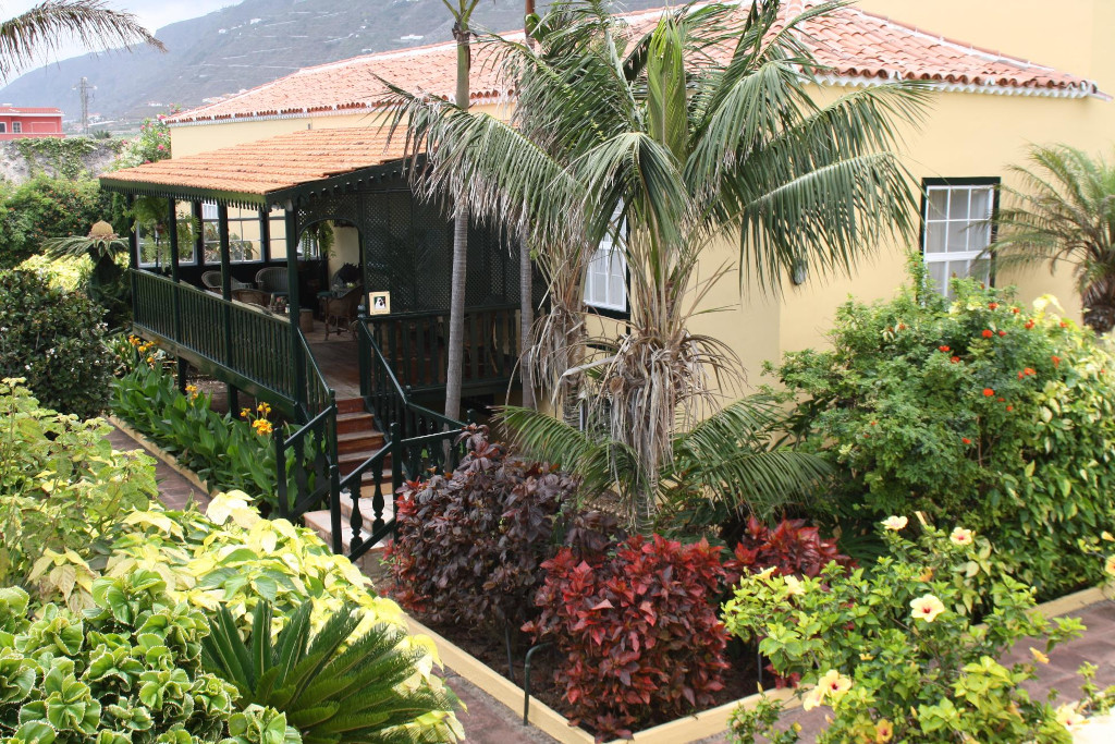Huis op Tenerife, omgeven door een tuin met palmbomen en kleurrijke struiken.