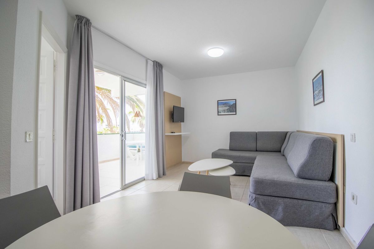 Woonkamer met een grijze bank en witte tafel, deur naar terras met palmbomen op Tenerife.