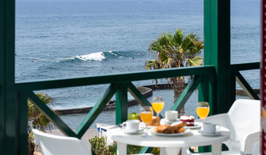 Ontbijt op een terras met uitzicht op zee op Tenerife, palmbomen en surfers op de achtergrond.