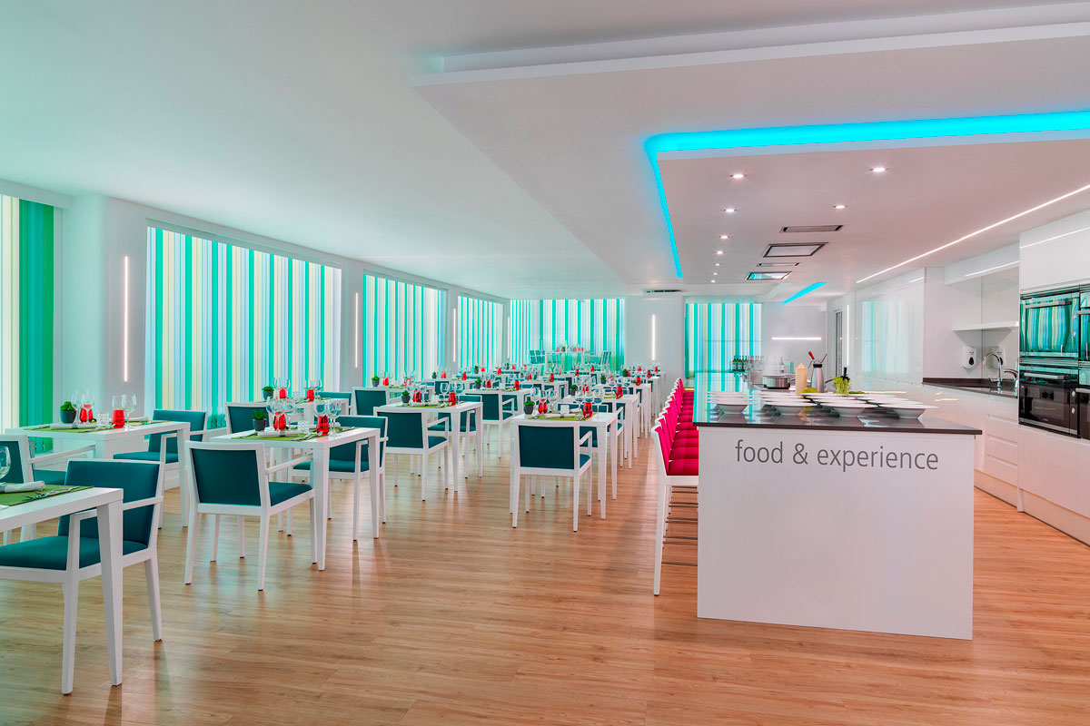 Restaurant verlicht met blauwe en roze stoelen, centrale tafel met eten en ervaring.