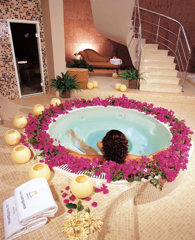 Persoon die ontspant in een bubbelbad omringd door bloemen, handdoeken met het label Les Meridien.