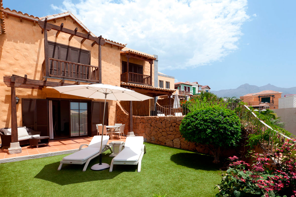 Huis met tuin op Tenerife, ligstoelen, parasol en planten op de voorgrond, heldere lucht.
