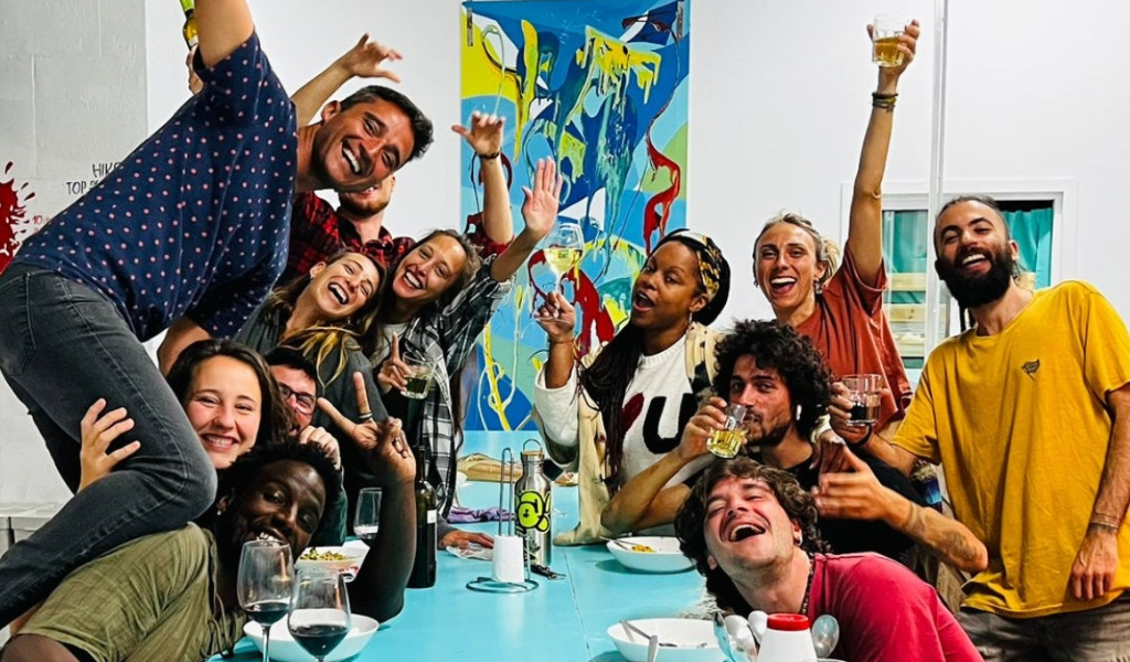 Groep vrienden die aan een tafel vieren met drankjes en een kleurrijke foto erachter.