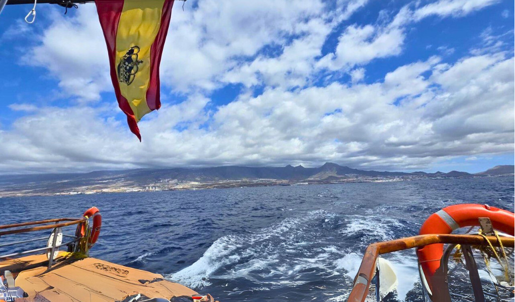 Zeilboot op zee met Spaanse vlag en uitzicht op de kust van Tenerife onder bewolkte luchten.
