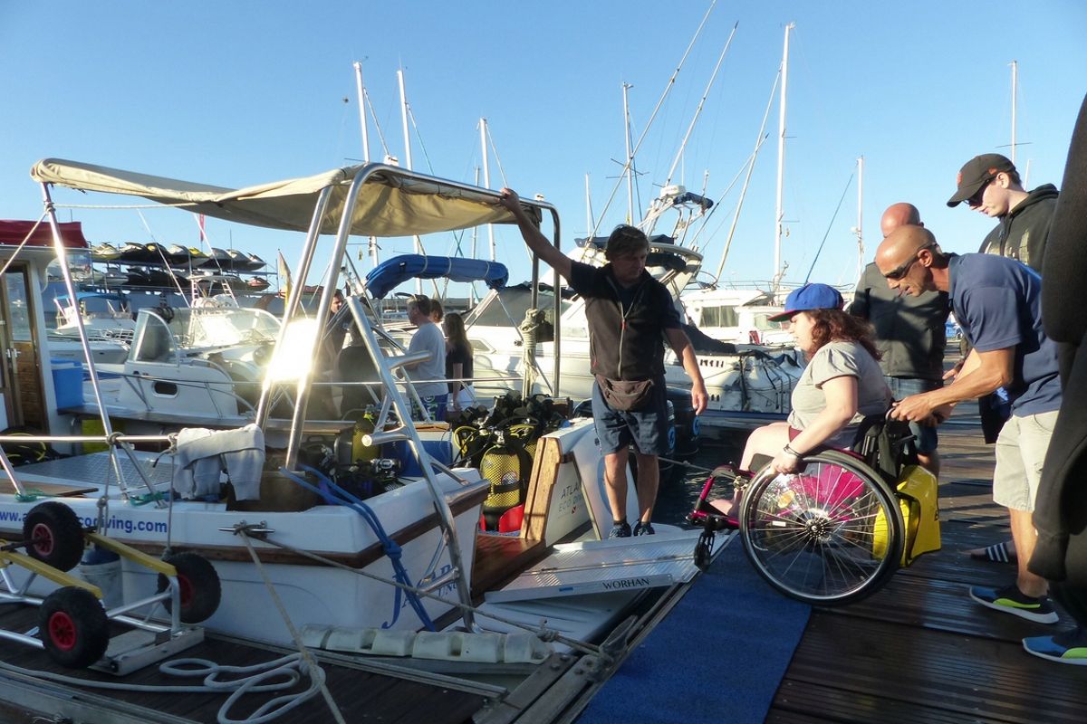 Persoon in een rolstoel die met hulp van meerdere mensen op Tenerife aan boord stapt van een boot.