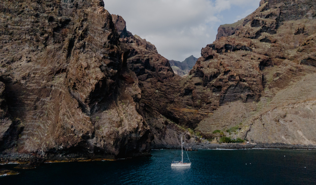 Zeilboot aan de rotsachtige kust van Tenerife met blauw water en bergen op de achtergrond.
