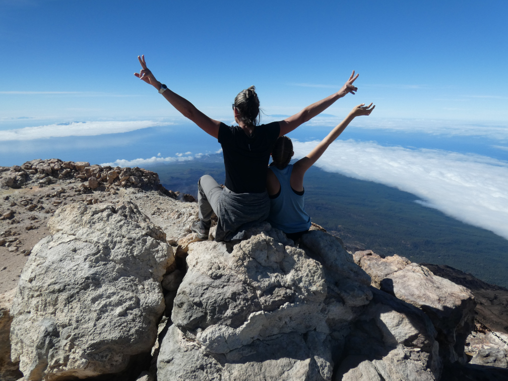 Mensen die hun armen omhoog steken op de top van de berg Teide op Tenerife, met uitzicht op het bewolkte landschap.