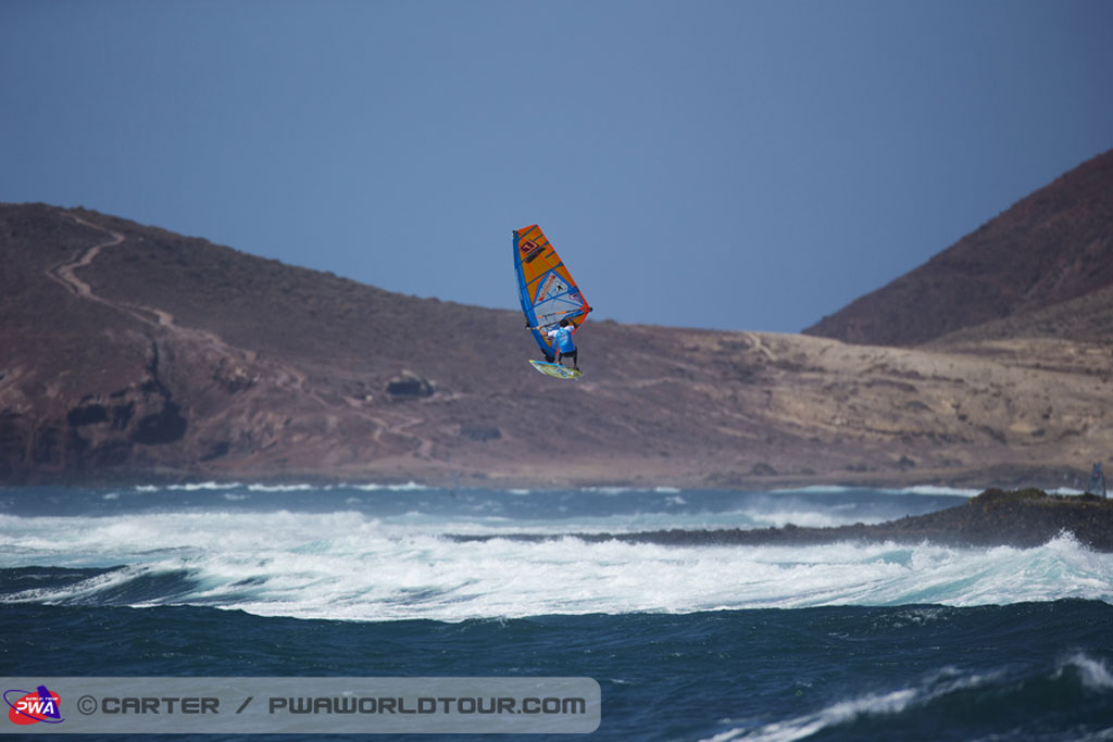 Windsurfer in de lucht in El Médano, Tenerife, met golven en bergen op de achtergrond.