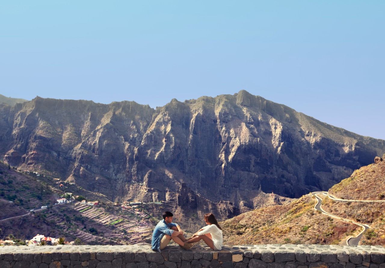 Mensen zitten voor de Masca-bergen op Tenerife, met een kronkelend pad op de achtergrond.